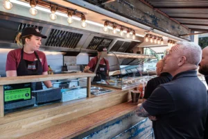 Catering staff in branded aprons serving a queue of guests from a mobile food unit at an outdoor corporate roadshow event