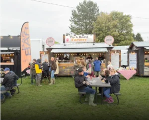 A "Pork Roast Carvery" food stall at an outdoor event, with people queuing and others eating at a picnic table on the grass.